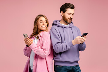 Smiling woman looking at smartphone of her bearded boyfriend isolated on pink background. Happy friends holding mobile phones, communication online. Technology, social media addiction concept