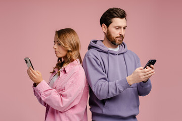 Couple of serious friends holding mobile phones, communication online isolated on pink background. Technology, social media addiction concept