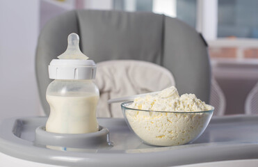 Fototapeta na wymiar A baby bottle with milk and a plate with dry milk formula enriched with bifidobacteria, vitamins and minerals stands on a gray high chair in the children's room. 