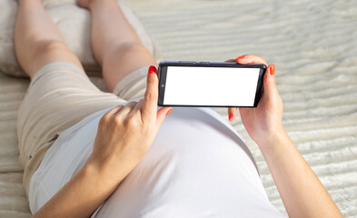 A pregnant girl lies on the bed and holds a smartphone in her hands. Concept of applications in...