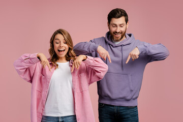 Happy couple wearing casual clothes isolated on pink background pointing fingers down on copy...