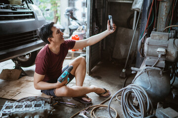 Young mechanic takes a selfie with cell phone while sitting with drill in garage