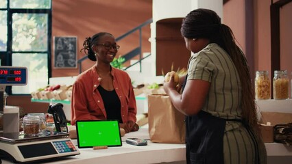 Customer approaching counter with greenscreen on device, buying eco friendly goods from local owner. African american vendor using isolated copyspace display on tablet, talks to client. Tripod shot.