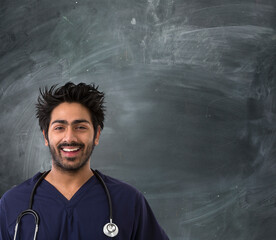 A happy Indian male doctor stands confidently in front of a blackboard. He wears navy scrubs and a...