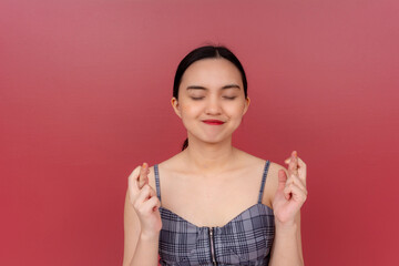 A hopeful young Asian lady stands with eyes closed and fingers crossed against a red background,...