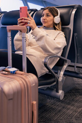Caucasian young woman sitting at the airport terminal while waiting for boarding for vacation. Uses smartphone for a video call or to listen music with a on carry-on suitcase next to her.