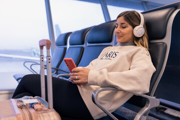 Caucasian young woman smiling and sitting at the airport terminal while waiting for boarding for vacation. Uses smartphone for a video call or to listen music with a on carry-on suitcase next to her.