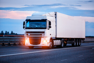 A truck with a refrigerated semi-trailer transports perishable food products in the evening against a blue sky. Import substitution, delivery of medicines, industry. Headlights on