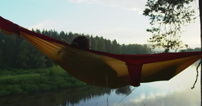 Man with dreadlocks from back swinging in a colorful hammock watching a river bend during dusk.