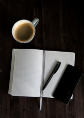 open white notebook , a black mobile phone and a cup of coffee on a dark wooden background