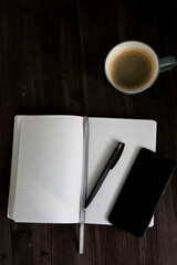 open white notebook , a black mobile phone and a cup of coffee on a dark wooden background