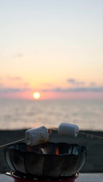 Vertical: Roast soft marshmallows on the fire of a portable gas burner on the sea coast at sunset using wooden arbecue skewers. It's romantic to make roasted marshmallows at sunset