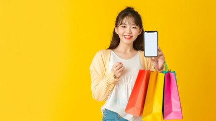 Mobile Shopping Application. Happy Asian Lady Showing Phone Blank White Screen Holding Colorful Paper Shopper Bags Standing Over Yellow Studio Background. Online Shopping Concept. Mockup   