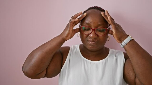 Stressed african american woman with braids, glasses, and plus size figure stands alone, wracked with headache ache over isolated pink background, wall of her struggles with anxiety, depression.