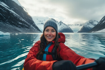 Young woman with a cute smile in red jacket and blue beanie kayaking amidst snowy mountains. Concept solo travelling, outdoor activities, adventure tourism.