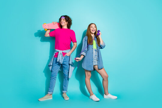 Full length photo of two teenagers boy hold skate and happy girl talking with vintage purple telephone isolated on cyan color background