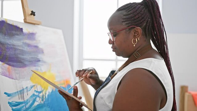 Focused african american woman artist, with braids and glasses, seriously drawing on canvas at indoor art studio