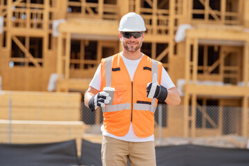 Coffee, engineer and construction worker relax on break at construction site. handsome male builder in hard hat smiling at camera. Construction Worker on Duty. Contractor and the Wooden House Frame