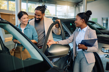 Happy multiracial couple communicating with saleswoman while buying new car in showroom.