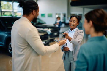 Happy black saleswoman giving to couple key of their new car in showroom.