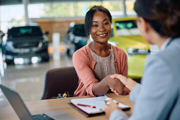 Happy black woman shaking hands with salesperson after buying new car in showroom.