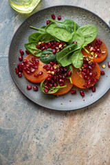 Plate with persimmon, fresh spinach and pomegranate salad on a beige granite background, vertical shot with space, high angle view