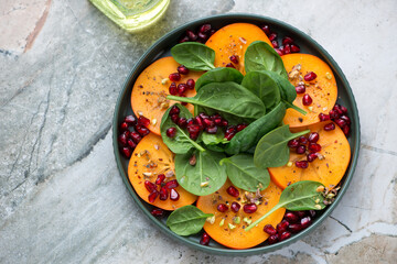 Plate with sliced persimmon, fresh baby spinach and pomegranate seeds, horizontal shot on a grey granite background, high angle view