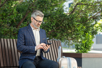 Fototapeta premium Mature man in a business suit using a phone while sitting on a bench outside an office with a suitcase, portraying a professional on the move.