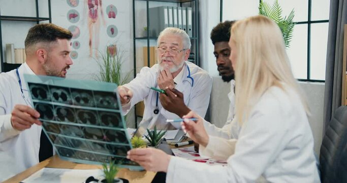Likable confident experienced diverse group of medics discussing patient's x-ray scan together with their respected bearded head doctor during joint briefing in workroom