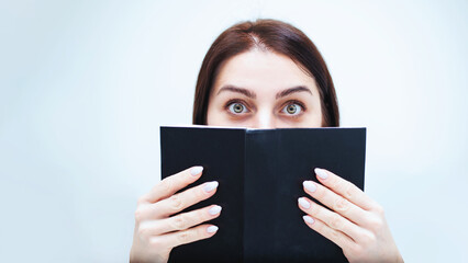 Young woman with tired eyes hides her face behind a black book on a white background