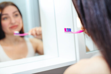 Beautiful woman in a white tank top holding a toothbrush with toothpaste near her mouth looking in the mirror in the bathroom at home - 719237732