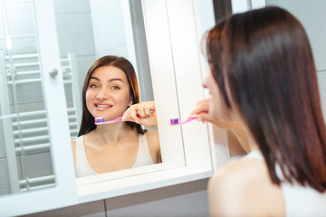 Beautiful woman in a white tank top holding a toothbrush with toothpaste near her mouth looking in the mirror in the bathroom at home - 719237762