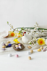 Easter still life. Colorful easter chocolate eggs in nest, spring flowers, chicken figurine composition on white wooden table. Space for text. Happy Easter! Seasons greetings