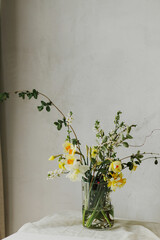 Beautiful daffodils, cherry bloom and greenery composition in glass vase. Stylish spring flowers bouquet on rustic table in rural room. Easter modern simple decor