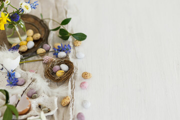 Fototapeta na wymiar Colorful easter chocolate eggs in nest, spring flowers, feathers and linen cloth on rustic wooden table. Space for text. Easter modern simple decoration. Happy Easter!
