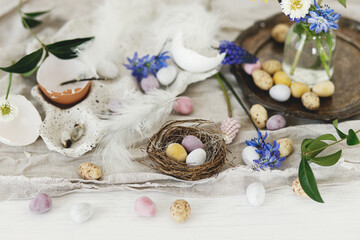 Fototapeta na wymiar Happy Easter! Stylish easter chocolate eggs in nest, spring flowers, feathers and linen cloth on rustic wooden table. Easter modern simple decoration still life