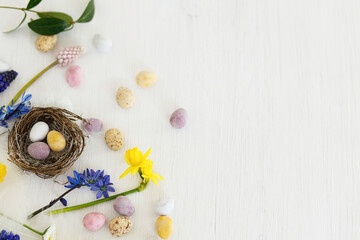 Easter rustic flat lay. Colorful easter chocolate eggs in nest, spring flowers, feathers border composition on white wooden table. Space for text. Happy Easter! Seasons greetings