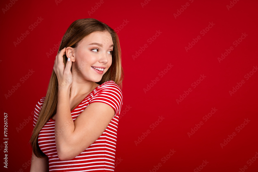 Poster Portrait of adorable teenager wear striped t-shirt tucking hair behind ear look at discount empty space isolated on red color background