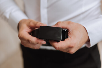 Close-up of a man holding a black leather belt. A man in a white shirt, without a face, holds a black leather belt in his hands. The morning of the groom at the wedding.