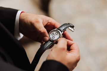 A man in a business suit holds a wristwatch in his hands, a mug of coffee is on the table in his...