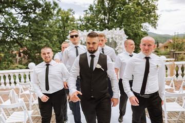 Cheerful, young, energetic witnesses of the groom next to the groom. Friends congratulate the groom. The groom in a vest and his friends in white shirts pose near the wedding arch. Wedding in nature.