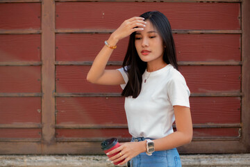 A teenage girl dressed in vintage clothing including a t-shirt and white pants stands alone holding a red coffee mug against the wall.