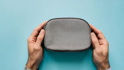 Young adult man hands holding and showing new gray cosmetic travel bag on light blue table background. Pastel color. Closeup. Point of view shot. Top down view - obrazy, fototapety, plakaty