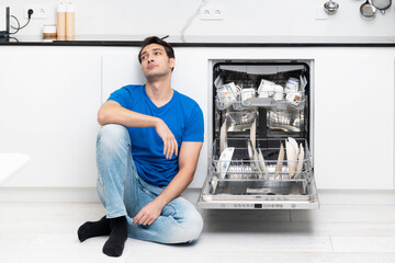 Tired and bored man putting dirty plates in dishwasher machine in the kitchen. Household and exhausting cleaning day concept