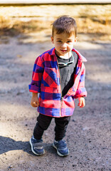 Cute little boy is standing on dirt road in red and black plaid jacket and black pants.