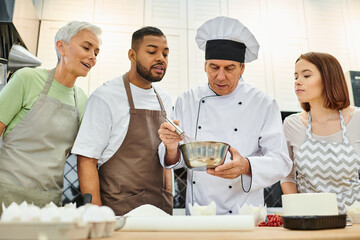 handsome mature chef in white hat showing how to whisk dough to diverse students, cooking courses