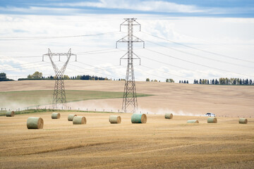 Round rolls on a harvested field overlooking distant utility provider electrical pylons on the Alberta prairies with a truck and dust trail on a gravel road in Rocky View Canada.
