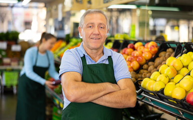 Portrait of confident mature male supermarket worker wearing apron standing in middle of grocery...