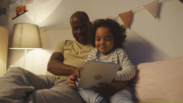Medium shot of African American dad sitting with baby son on bed and hugging him while playing, watching or reading on tablet in cozy modern bedroom