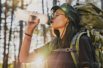 Profile photo of young woman drinking water in forest, sun flare. Female explorer traveler adventurer rehydrating with cold drink, relaxing feeling thirsty in solo trip - obrazy, fototapety, plakaty